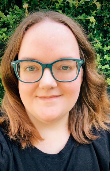 An attractive young person of European descent with light brown hair, a black t-shirt, and large square glasses the same blue-green color as their eyes. They're facing the camera with a friendly and calm smile that says, "I'm a laid-back person who's great